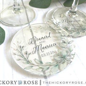 May include: Round glass coaster with a watercolor design of green leaves and white flowers. The coaster has the text "Diamond and Mauricio 03.21.26" printed on it.
