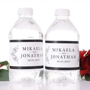May include: Two clear plastic water bottles with white caps and labels. The labels read "MIKAELA AND JONATHAN 08.01.2019" in black text with decorative flourishes. A red rose is visible in the background.