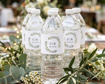 Op de afbeelding: Doorzichtige plastic waterflessen met witte etiketten voorzien van een groen botanisch kransontwerp. De tekst op de etiketten bevat de initialen C|R en de datum 06.12.24. De flessen staan opgesteld op een houten ondergrond.