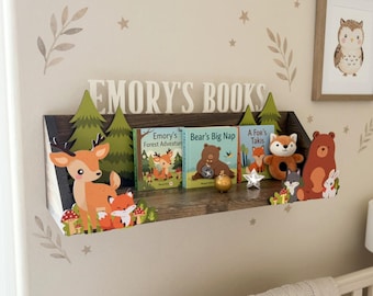 Op de afbeelding: Een houten plank met de tekst "EMORY'S BOOKS" toont kinderboeken en decoraties met bos thema. De plank bevat een hert, een vos, een beer en andere bos elementen. Een ingelijste uil afbeelding hangt aan de muur.