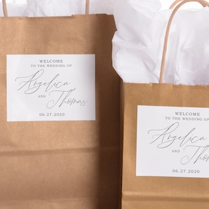 May include: Two brown paper gift bags with white tissue paper inside. Each bag has a white sticker with the text "Welcome to the wedding of Angelica and Thomas 06.27.2020".