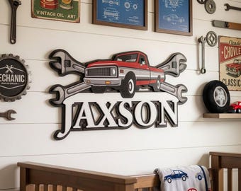 Op de afbeelding: Een wanddecoratie met een ontwerp van een rode en grijze pick-up, gekruiste moersleutels en de naam "JAXSON" in grote letters. De muur is versierd met kunst en gereedschap met een vintage autothema, waardoor een garage- of automotief thema ontstaat.