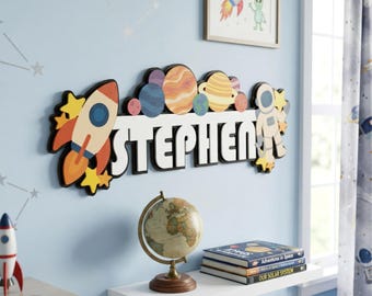 Op de afbeelding: Een houten naambord met de naam "STEPHEN" in witte blokletters, versierd met een raket, planeten en een astronaut. Het bord is bevestigd aan een lichtblauwe muur, met een ingelijste tekening van een raket en een alien.