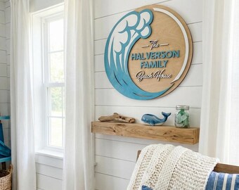 Op de afbeelding: Een rond houten bord met een blauw golfontwerp en de tekst "The Halverson Family Beach House". Het bord is bevestigd aan een witte lambrisering. Een houten plank eronder bevat een walvisbeeldje en een pot.
