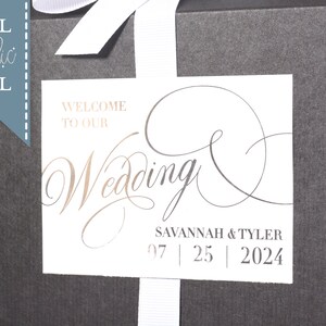 May include: A white wedding favor box with a white ribbon and a white label with the text "Welcome to our Wedding" in gold foil lettering. The label also includes the names "Savannah & Tyler" and the date "07 | 25 | 2024".