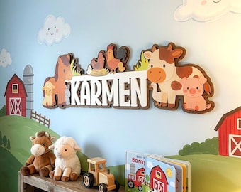 Op de afbeelding: Een houten wanddecoratie met boerderijthema met de naam "Karmen" in witte letters. Het ontwerp omvat een paard, schaap, koe, varken en andere boerderijdieren. Een houten plank eronder bevat knuffels, een speelgoedtrekker en een boek.