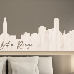 May include: A white wooden cutout of the Baton Rouge skyline with the city name written below. The cutout is mounted on a light brown wall above a white headboard.