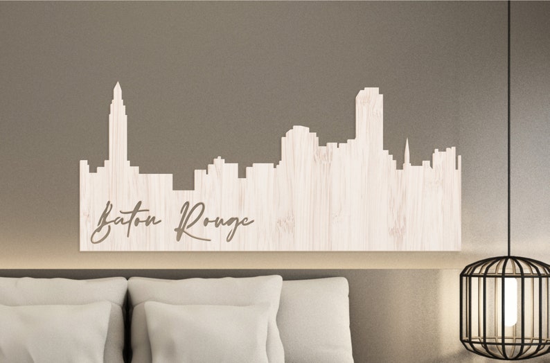 May include: A white wooden cutout of the Baton Rouge skyline with the city name written below. The cutout is mounted on a light brown wall above a white headboard.
