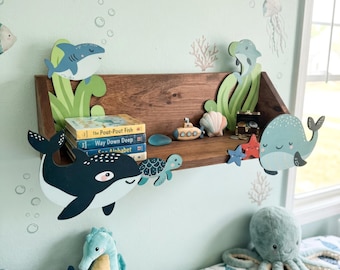 Op de afbeelding: Een houten plank met decoraties met oceaanthema en boeken. De plank heeft uitsparingen van een walvis, haai en andere zeedieren. Boeken zijn onder meer "The Pout-Pout Fish" en "Way Down Deep". De muur is lichtblauw geschilderd.