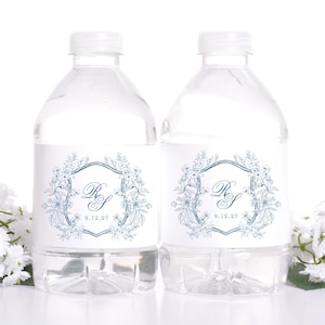May include: Two clear water bottles with white caps. Each bottle has a white label with a blue floral design, the initials "RS", and the date "9.12.27". The bottles are set against a white background.