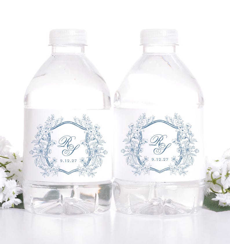 May include: Two clear water bottles with white caps. Each bottle has a white label with a blue floral design, the initials "RS", and the date "9.12.27". The bottles are set against a white background.