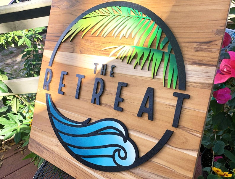 May include: A wooden sign with a circular design featuring a palm tree, a wave, and the text "THE RETREAT" in black lettering.