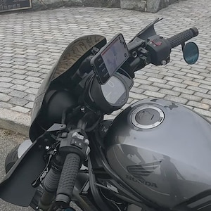 Könnte beinhalten: Ein dunkelgraues Motorrad mit Windschutzscheibe, einem am Lenker montierten Telefon und einem Seitenspiegel. Das Honda-Logo ist auf dem Kraftstofftank zu sehen. Die Lenker haben strukturierte Griffe.