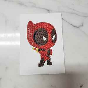 Puede incluir: Una pegatina con el personaje de dibujos animados Deadpool. El personaje está representado en una pose de pie, con una combinación de colores rojo y negro. La pegatina está cubierta de pequeños adornos redondos rojos y marrones.