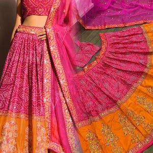 May include: A vibrant pink and orange traditional Indian outfit. The ensemble includes a crop top, a long skirt, and a sheer scarf. The skirt features intricate embroidery and a contrasting orange hem. The outfit is adorned with gold accents and detailed patterns.