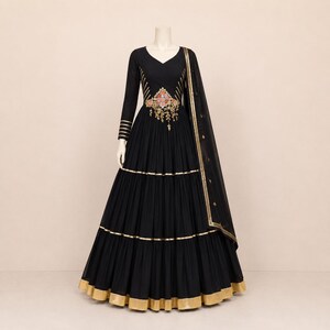 May include: A black floor-length gown with long sleeves, featuring a V-neckline and a central waist panel with colorful floral embroidery. The skirt has horizontal gold bands, and a matching black sheer dupatta with a gold border is draped.