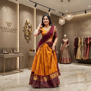 May include: A two-piece ensemble consisting of a mustard yellow pleated skirt and a matching short-sleeved blouse, draped with a deep maroon shawl featuring intricate gold border detailing. The outfit is worn in a retail space with Westland Clothing signage.