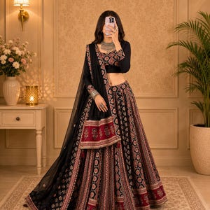 Puede incluir: Un conjunto de lehenga choli negro que presenta una blusa de manga larga y una falda hasta el suelo con intrincados patrones florales en rojo y blanco. El conjunto incluye una dupatta negra transparente con un borde ancho estampado en tonos rojos y blancos.