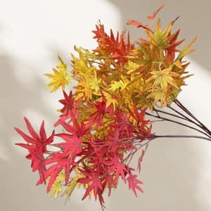 May include: A bundle of artificial maple leaves in vibrant autumn colors. The leaves transition from deep red to golden yellow, creating a realistic and decorative display. The stems are dark brown.
