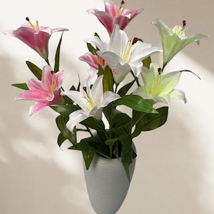 May include: A vase of lilies in a white vase. The lilies are in shades of pink, white, and light green, with dark green leaves. The vase is a light grey colour and has a textured surface. The flowers are arranged in a full, lush display.