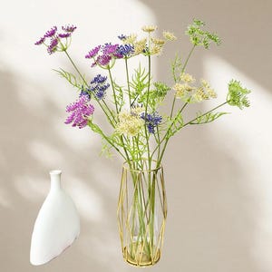 May include: A bouquet of colorful artificial flowers in a gold geometric vase. The flowers include purple, blue, and cream-colored blooms with green stems and leaves. A white, teardrop-shaped vase sits to the left.