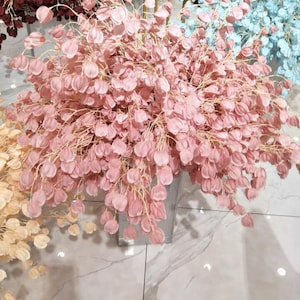 Può includere: Un bouquet di fiori rosa artificiali con delicate forme a petalo. I fiori sono disposti in un contenitore di metallo argentato, creando un'estetica morbida e romantica. Gli steli sono di colore beige chiaro. Altri bouquet sullo sfondo sono blu e gialli.