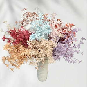 Può includere: Un vivace bouquet di fiori artificiali in un vaso di ceramica bianco. I fiori presentano una varietà di colori, tra cui rosso, arancione, blu, viola e crema. La composizione è piena e crea un'esposizione visivamente accattivante.