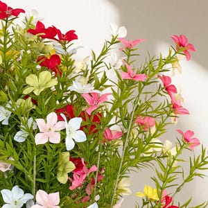 May include: A colourful arrangement of artificial flowers in shades of red, pink, white, yellow, and green. The flowers feature delicate petals and are arranged with green stems and leaves. The display is set against a white backdrop.