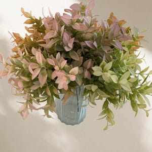 May include: A bouquet of artificial greenery in a light blue vase. The leaves display a mix of colors, including light green, pink, and a touch of orange. The vase has a textured surface and is set against a neutral background.