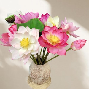 Op de afbeelding: Een boeket kunstmatige lotusbloemen in verschillende tinten roze, wit en geel, met groene stelen en bladeren. De bloemen zijn gerangschikt in een getextureerde, beige vaas, op een geweven mat.