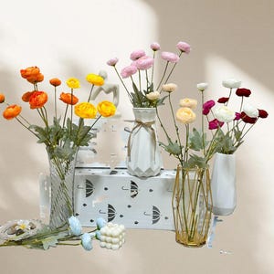 May include: A display of artificial flowers in vases of different styles. The flowers are in shades of orange, yellow, pink, and white. The vases are clear, white, and gold, set on a surface with an umbrella design.