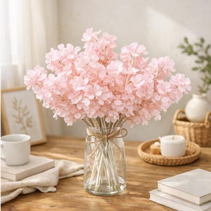 May include: A bouquet of light pink artificial flowers in a clear glass vase tied with twine. The flowers have multiple delicate petals. The vase sits on a wooden table with other decorative items, including books and a white mug.