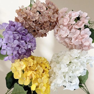 May include: A selection of artificial hydrangea flower bouquets in shades of purple, yellow, white, pink, and brown. Each bouquet has multiple flower heads and green leaves, offering a colourful and natural appearance.