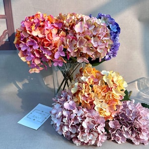 以下が含まれることがあります： ピンク、オレンジ、イエロー、パープルの色合いの人工アジサイの花のコレクション。1つの束は透明なガラスの花瓶に生けられ、他の束は近くに置かれています。LOVEという文字が書かれた小さな白い長方形のカードが見えます。