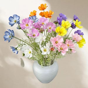 May include: A vibrant bouquet of cosmos flowers in a white vase. The flowers are in shades of blue, pink, yellow, orange, and white. The vase is light grey with a textured surface. The arrangement is set against a neutral background.