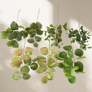 May include: Three artificial eucalyptus branches with varying shades of green and yellow-green leaves. Some leaves have red veining. The branches are suspended against a neutral background, showcasing the different leaf shapes and colors.