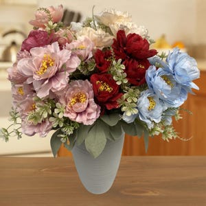Puede incluir: Un ramo de flores artificiales en un jarrón gris claro. El arreglo incluye peonías rosas, rojas y azules, con follaje verde. El jarrón se encuentra sobre una superficie de madera, creando una vibrante exhibición floral.