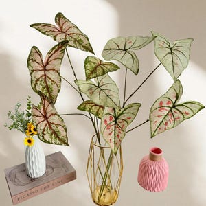 Puede incluir: Hojas artificiales de Caladium con patrones rosas y verdes se exhiben en un jarrón geométrico dorado. También hay un jarrón blanco pequeño con flores amarillas y un jarrón rosa, junto con un libro titulado "Picasso The Line."