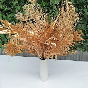 Puede incluir: Un ramo de plantas artificiales doradas en un jarrón de cerámica blanco. El arreglo incluye varias texturas de hojas y helechos, creando una exhibición completa y decorativa. El fondo presenta follaje verde.