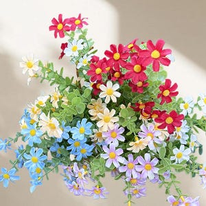 Peut inclure: Un arrangement floral artificiel aux couleurs vives. Le bouquet présente des fleurs en forme de marguerites rouges, blanches, bleues, jaunes et violettes avec des centres jaunes, complétées par un feuillage vert. Un accent floral décoratif.