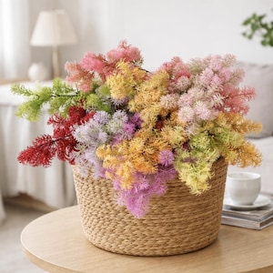 May include: A wicker basket overflowing with vibrant artificial flowers. The flowers display shades of red, pink, yellow, orange, green, and purple. The basket rests on a round wooden table. A white cup and saucer are also on the table.