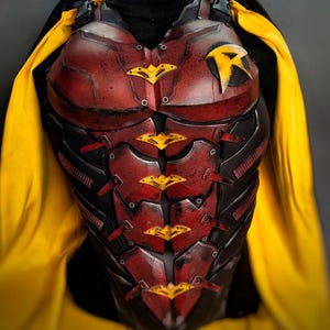 Può includere: Un pettorale corazzato rosso e nero con un emblema R giallo sul lato sinistro e accenti gialli a forma di pipistrello al centro. L'armatura è indossata con un dolcevita nero e un mantello giallo brillante che ricade sulle spalle.