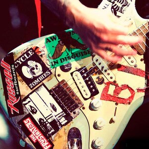 May include: A light blue electric guitar covered in stickers, including "Russell Surfboards," "Zombies," and "The Frustrators." A hand is strumming the strings. The guitar has white knobs and a wooden neck.