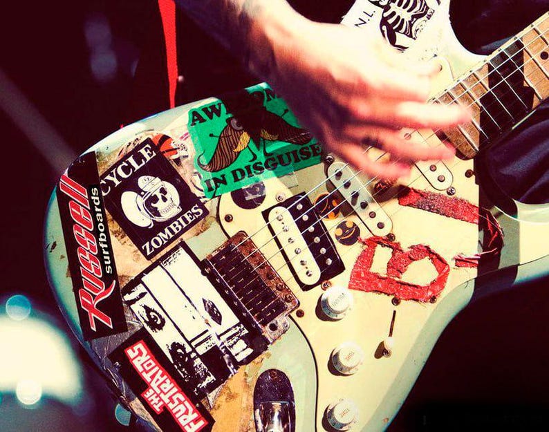 May include: A light blue electric guitar covered in stickers, including "Russell Surfboards," "Zombies," and "The Frustrators." A hand is strumming the strings. The guitar has white knobs and a wooden neck.