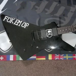 May include: Black electric guitar with the text "FOK EM UP" in white. The guitar features a skull and crossbones emblem. The instrument is set against a backdrop with a flag design.