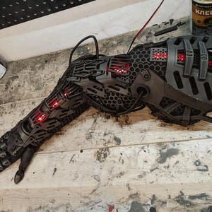 May include: A dark grey robotic arm with red internal lighting, showcasing a complex, layered design with hexagonal patterns and articulated sections. Wires and cables extend from the arm, suggesting a technological or sci-fi theme. The arm is likely a prop or a piece of art.
