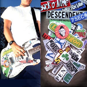May include: An electric guitar covered in stickers, held by a person in a white t-shirt. A collection of stickers with band names and graphics is displayed on a brown surface.