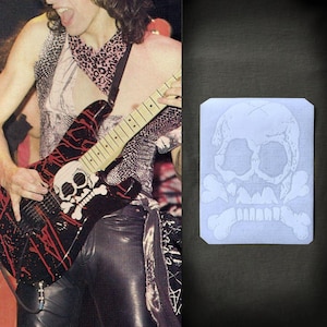 May include: Black electric guitar with red splatter design and a skull and crossbones graphic. The guitar is being played. A skull and crossbones design is also on a white rectangle.