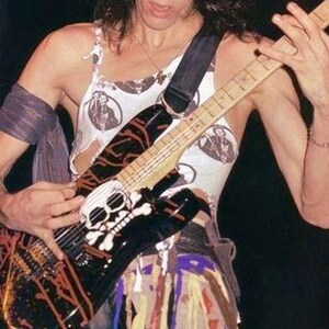 May include: A black electric guitar with a skull and crossbones design, orange accents, and a white pickguard. The musician wears a white tank top with circular black graphics and a strap. The guitar is being played.