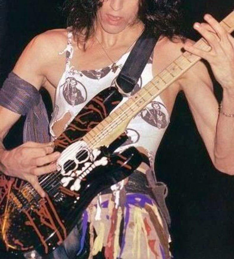 May include: A black electric guitar with a skull and crossbones design, orange accents, and a white pickguard. The musician wears a white tank top with circular black graphics and a strap. The guitar is being played.
